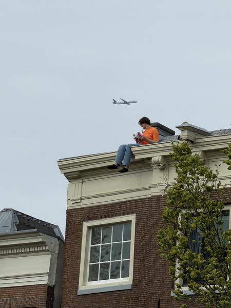 A person sits calmly on the ornate rooftop of a classic Amsterdam canal house, engrossed in their phone or a book, while an airplane flies high in the overcast sky